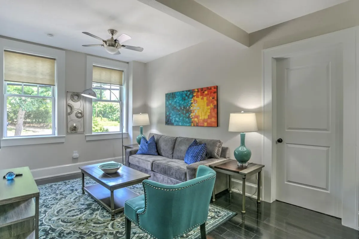 This image captures a welcoming living area featuring a comfortable gray sofa, vibrant artwork on the walls, a teal accent chair, and warm table lamps. The room is bathed in natural light, highlighting the blend of plush furnishings and thoughtful decor.