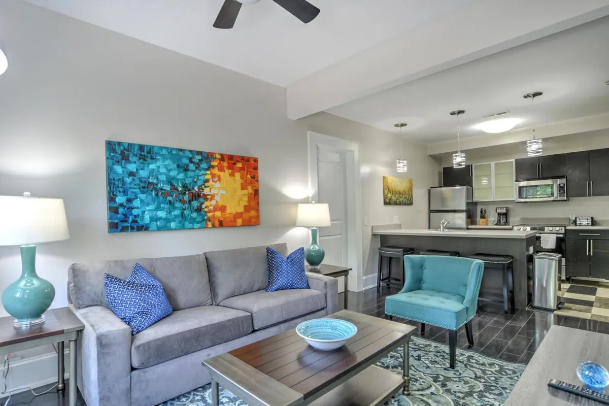 This inviting living area features a plush sofa, striking blue armchair, and a well-lit kitchen area in the background. Artwork adorns the walls, and a coffee table sits at the room’s center, suggesting a comfortable space for relaxation or informal meetings. The open layout hints at a blend of work and leisure, making it ideal for business travelers seeking both productivity and downtime.