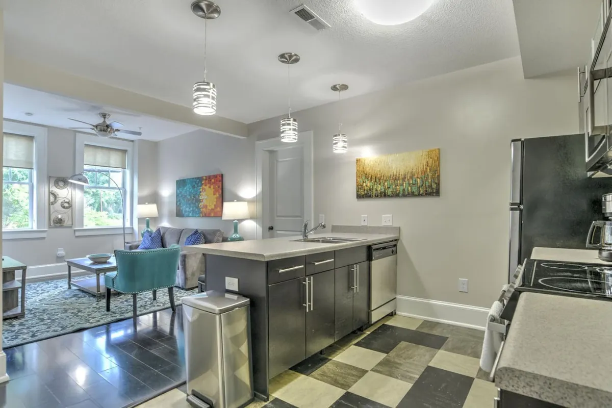 The image showcases a bright, inviting open-concept living space within the historic King James Building. A stylish kitchen island takes center stage, complemented by vibrant, local artwork and a plush seating area framed by large, sunlit windows. The scene evokes a contemporary yet cozy atmosphere, perfect for cooking, relaxing, or socializing.