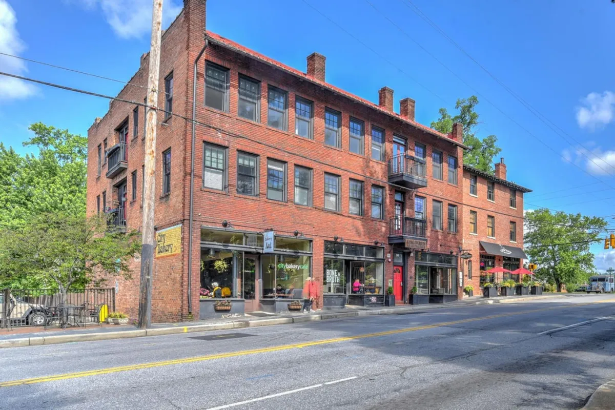 A brick facade with large windows and a welcoming balcony, flanked by shopfronts where people are shopping, dining outdoors, and socializing. The atmosphere is bustling, highlighting the presence of unique local businesses and community events.