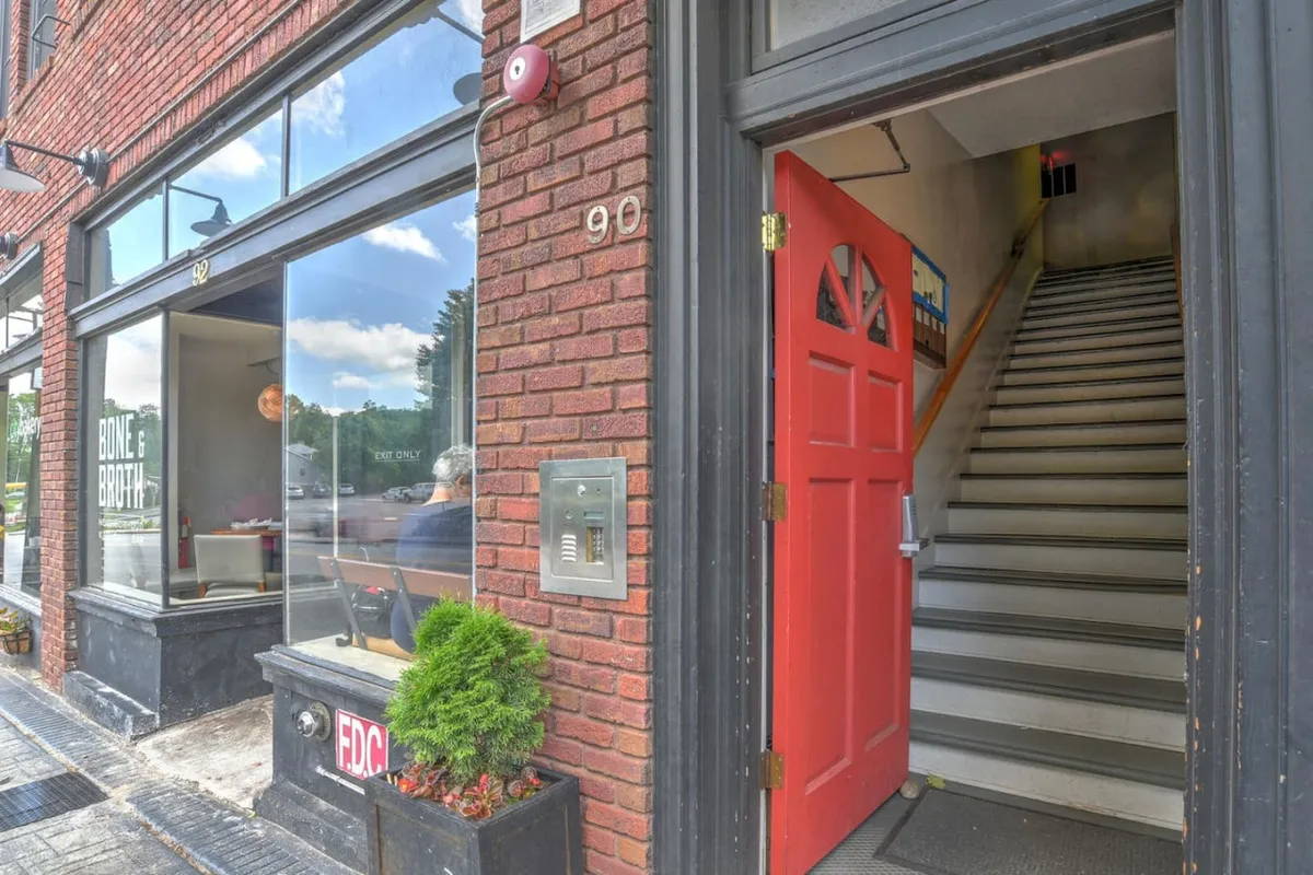 The image features the exterior of a historic brick building with a striking red door, large windows, and a welcoming staircase. The street-level view captures vibrant outdoor activity, with people visiting a cafe, exploring neighboring shops, and socializing in the inviting atmosphere. The setting suggests a lively and well-preserved district, reflecting Asheville’s unique architectural character.