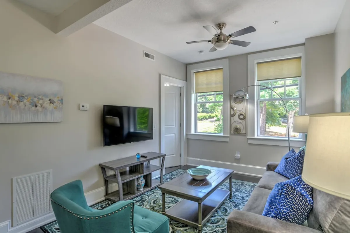A cozy living space is pictured with a sofa sporting blue cushions, a contemporary coffee table, a TV, and a window that lets in ample daylight. A decorative painting above the sofa and the uncluttered, welcoming layout reflect a harmonious blend of comfort and style.