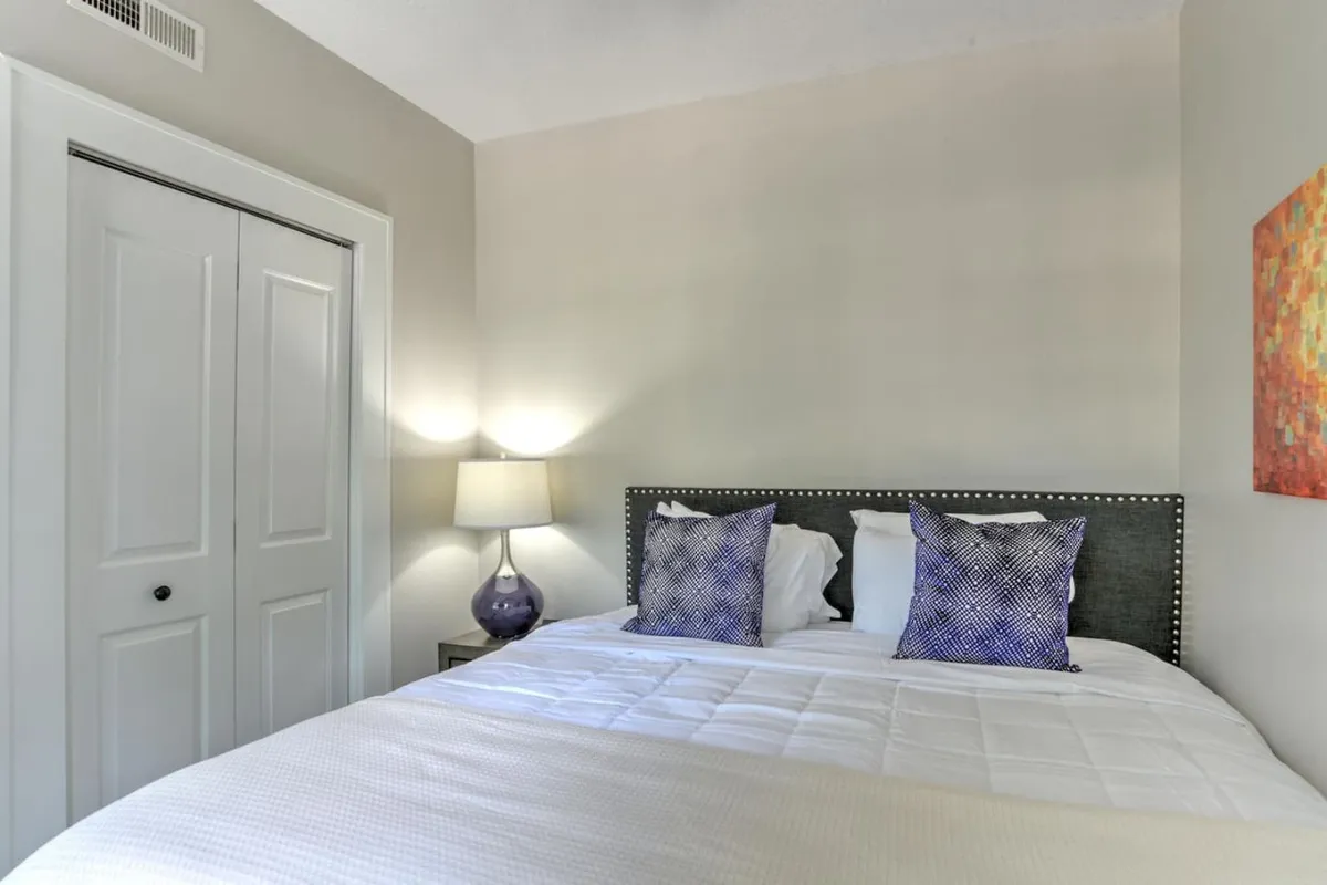 A calm, cozy bedroom showcasing a stylish bed with decorative pillows, a softly illuminated table lamp, and art hanging on the wall. The space feels inviting, perfect for relaxing after a busy day.