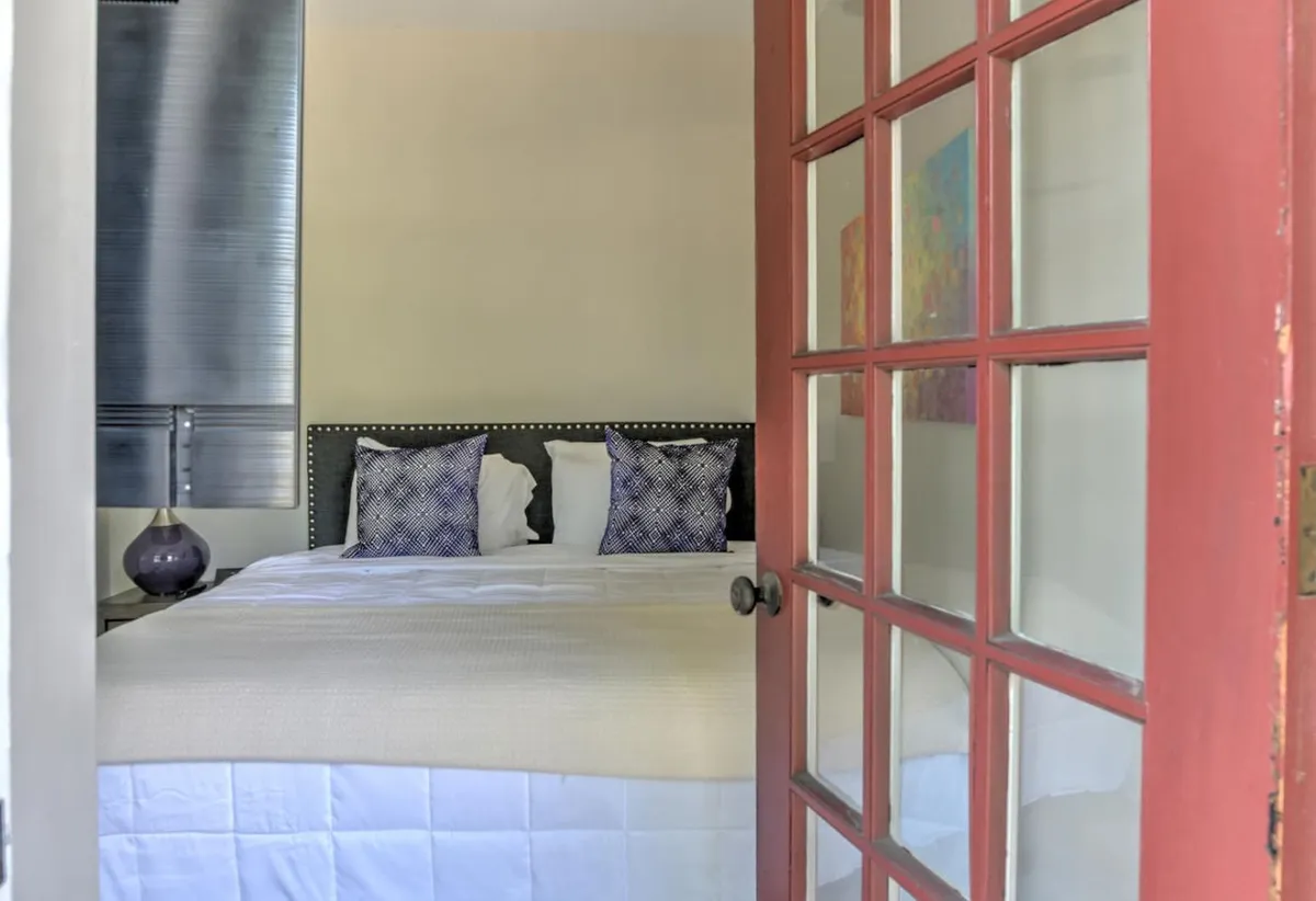 A cozy bedroom with a bed decorated in patterned pillows, a bedside table with a vase, and colorful artwork above the headboard. Sunlight streams in to create a restful atmosphere—ideal for unwinding after a busy day exploring the neighborhood or working downtown.