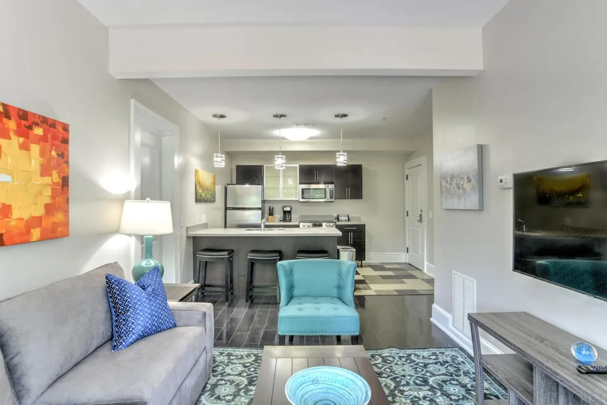 The image presents a spacious open-plan area featuring a plush sofa, a modern turquoise chair, a functional kitchen island, and wall art. The arrangement encourages relaxation and socializing, while the natural light and tasteful decor underline the building’s historic-meets-modern ambiance.