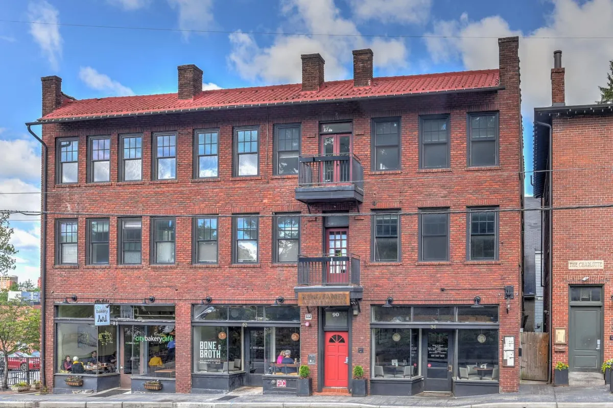 A vibrant exterior view featuring a red door, second-floor balcony, and large windows, with visible storefronts hinting at local cafes and boutiques. People can be seen dining at nearby cafes, shopping at local boutiques, and casually observing from the balcony, giving life and energy to the scene.