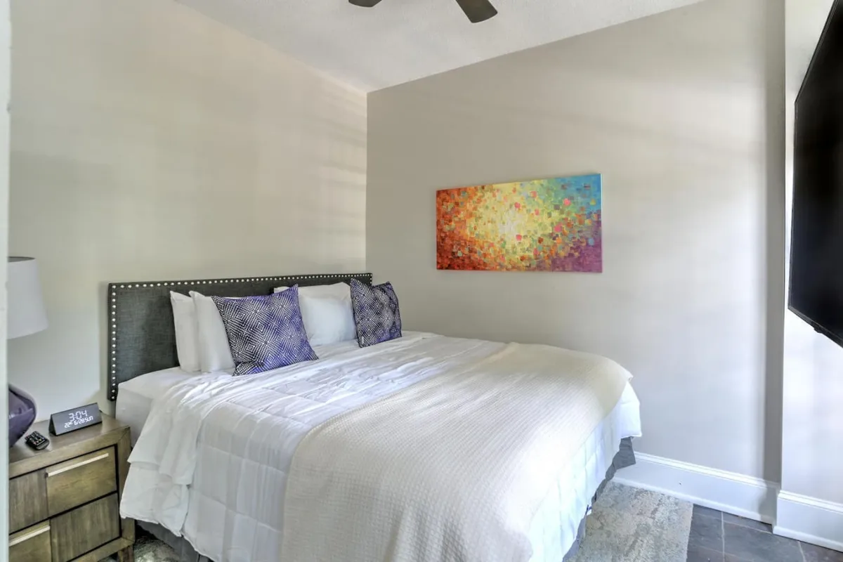A tranquil bedroom scene featuring a comfortable bed adorned with decorative pillows, colorful wall art above the headboard, and a nightstand with a soft-glowing lamp. Morning light filters in, giving the space a peaceful and cozy feel.